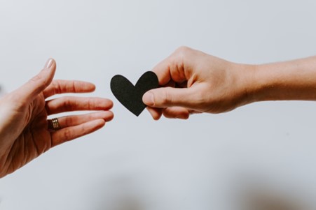 Image shows two hands, one passing a black cut out paper heart to the other hand.